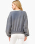 A woman shown from the back wearing a faded slate blue-gray crewneck sweatshirt and white pants, photographed against a clean white background.