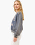 A woman modeling a washed slate blue cropped sweatshirt with cream collegiate-style lettering, paired with white pants, against a plain white studio background.