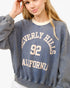 A woman wearing a faded blue-gray crewneck sweatshirt with 'BEVERLY HILLS 92 CALIFORNIA' printed in cream varsity lettering on the front.