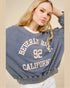 Person wearing a blue 'Beverly Hills 92 California' sweatshirt against a beige background