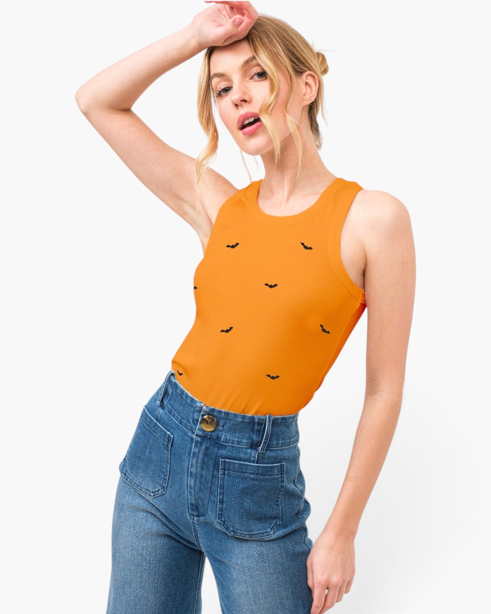 A woman modeling a bright orange sleeveless crop top with small black bat motifs, paired with high-waisted blue jeans, against a plain white studio background.