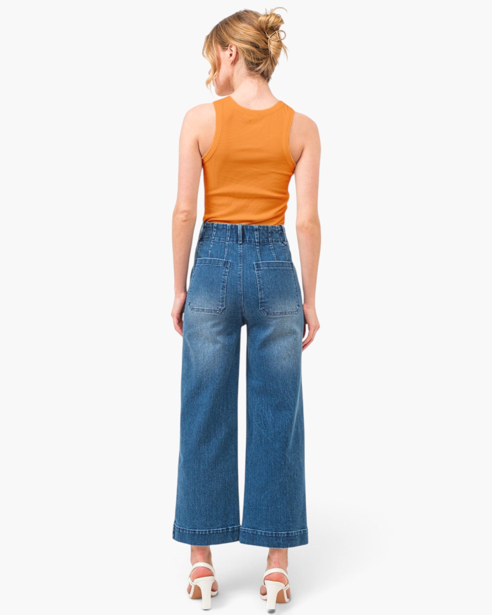 A woman shown from the back wearing a bright orange sleeveless top and high-waisted blue wide-leg denim jeans, paired with white heeled sandals, against a plain white studio background.