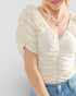 Woman wearing a cream-colored ruched top with short sleeves against a light gray background