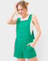 A woman wearing a bright green sleeveless short romper with front buttons and side pockets, layered over a white tank top, against a light gray studio background.