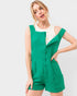 A woman modeling a bright green textured sleeveless romper with front buttons and side pockets, worn over a white top, against a light gray studio background.
