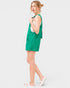 A woman modeling a bright green sleeveless overall-style romper with front patch pockets, worn over a white tank top and white slide sandals, against a light gray studio background.