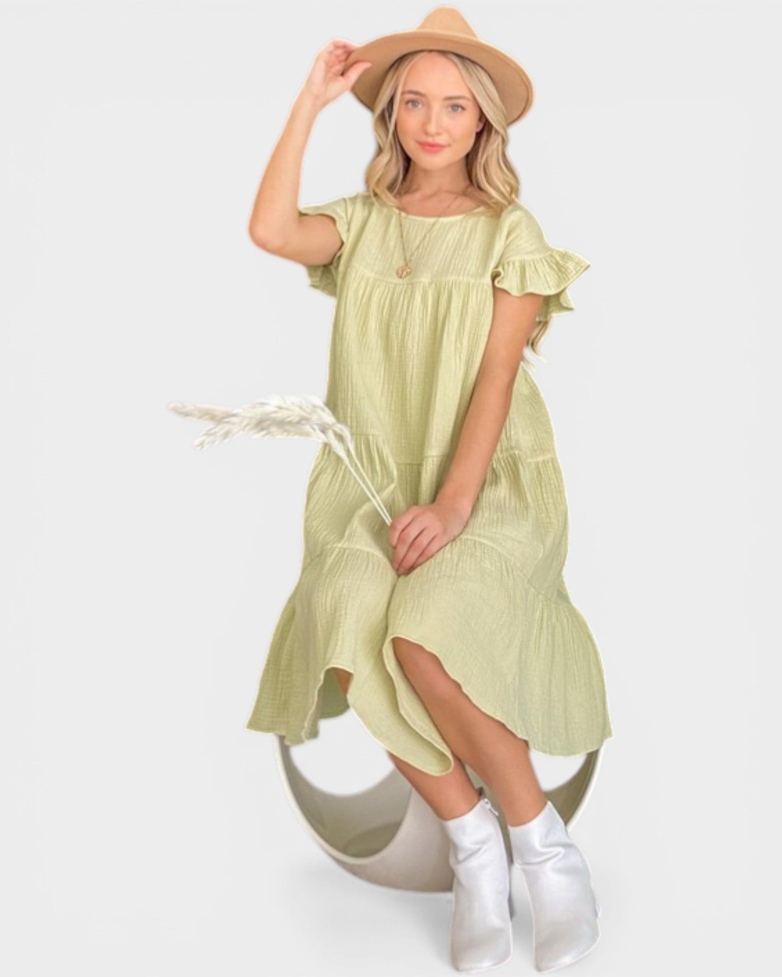 Woman wearing a light green dress with ruffled sleeves and a hat, holding a dried flower on a white background.