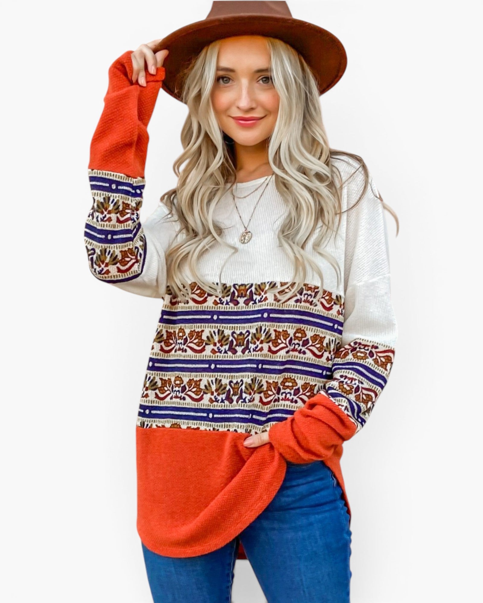 A woman wearing a long-sleeve color-block knit sweater with a white upper section, a multicolor patterned middle band, and a bright orange lower section and cuffs, paired with blue jeans and a brown hat against a white background.