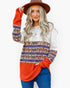 A woman wearing a long-sleeve color-block knit sweater with a white upper section, a multicolor patterned middle band, and a bright orange lower section and cuffs, paired with blue jeans and a brown hat against a white background.