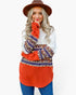 A woman wearing a bohemian-style knit tunic sweater with a white long-sleeve top underneath, featuring a bright orange hem and cuffs with intricate brown, blue, and white patterns. She is also wearing a brown wide-brim hat and blue jeans, posed against a solid white background.