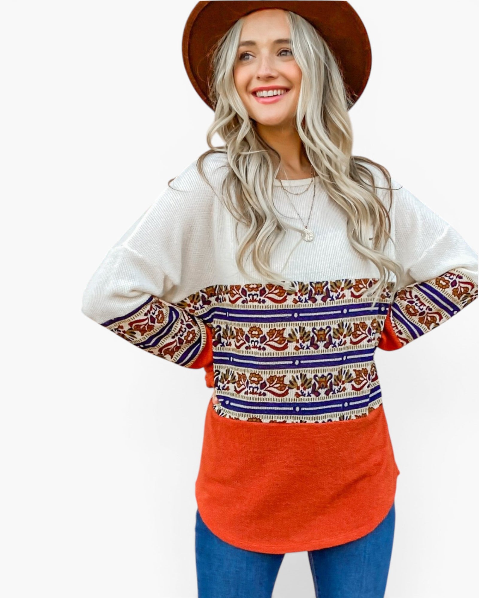 A woman wearing a long-sleeve crewneck sweater with a color-blocked design: off-white on top, a multicolor bohemian pattern band across the chest and sleeves, and a bright orange-red lower section, paired with blue jeans and a brown hat against a white background.