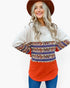 A woman wearing a long-sleeve crewneck sweater with a color-blocked design: off-white on top, a multicolor bohemian pattern band across the chest and sleeves, and a bright orange-red lower section, paired with blue jeans and a brown hat against a white background.