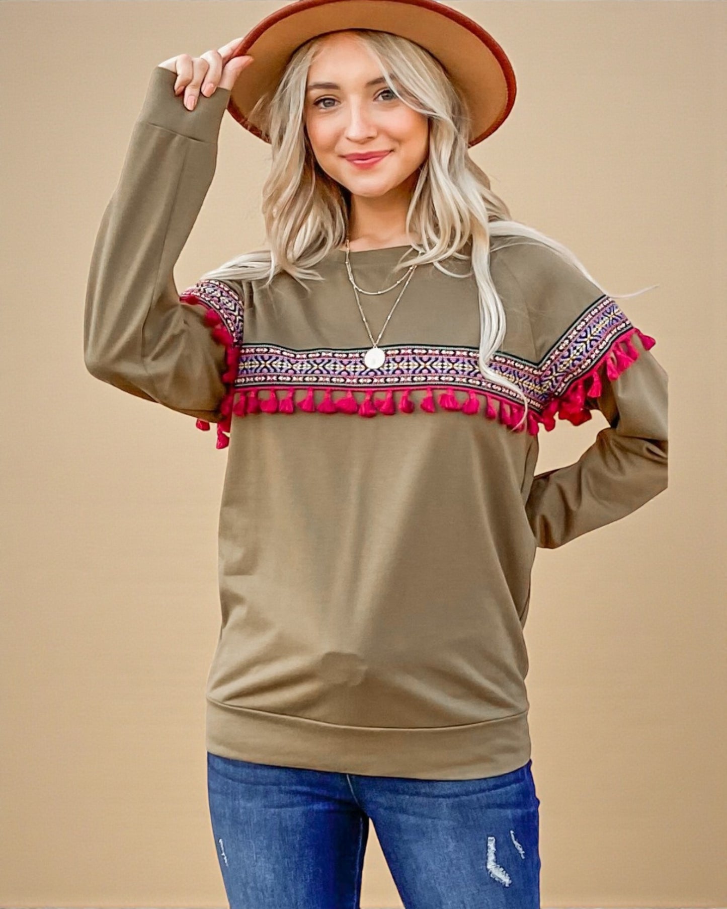 Woman wearing a brown sweater with red and white patterned trim, blue jeans, and a beige hat on a beige background.