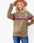 A woman wearing a long-sleeve crewneck sweatshirt in olive brown, decorated with a colorful geometric pattern band and bright pink tassels across the shoulders. She is also wearing a brown wide-brim hat and blue jeans, posing against a solid white background.