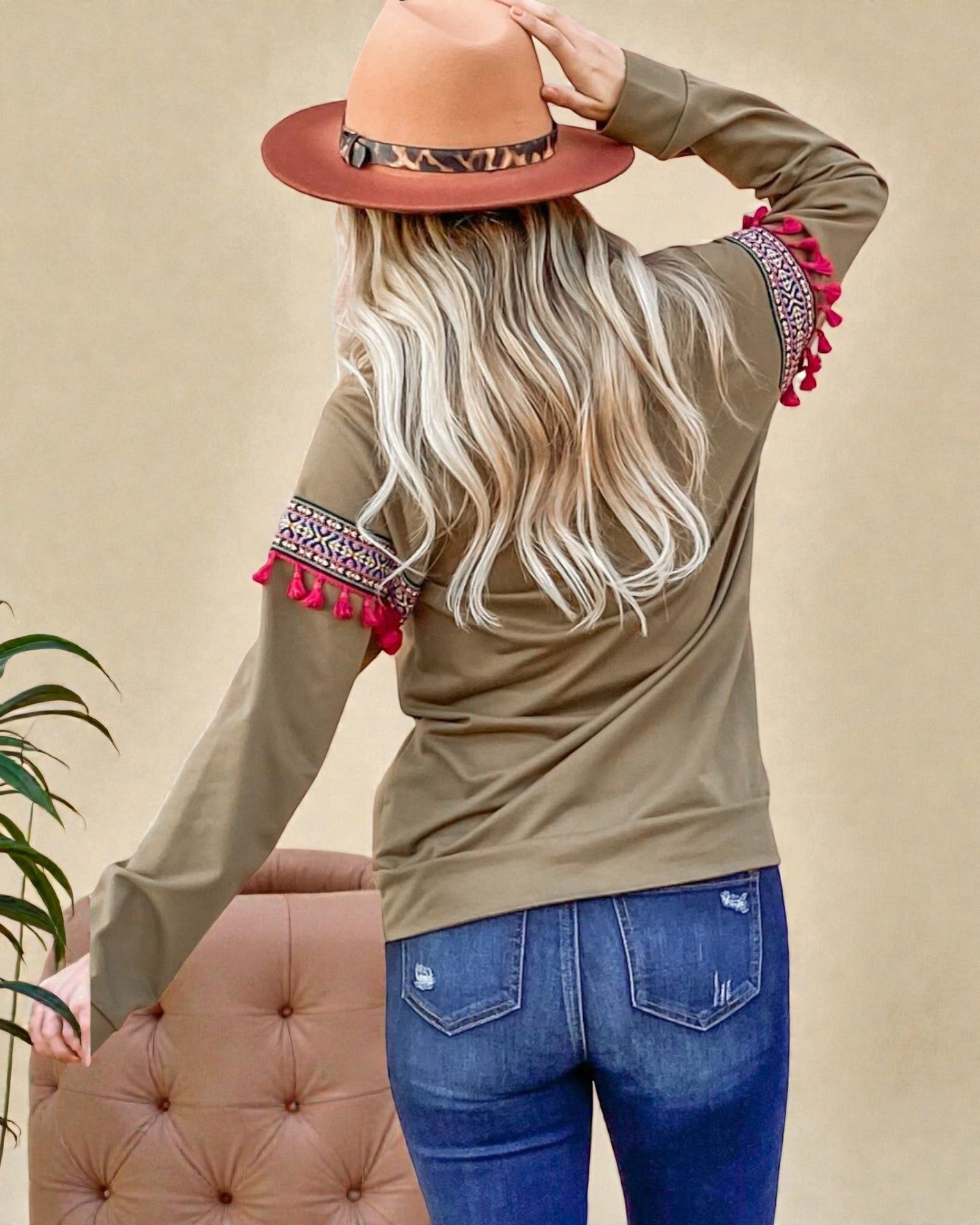 Person wearing a beige long-sleeve top with red tassel details and blue jeans, standing against a beige background.