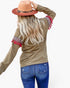 A woman shown from the back wearing a long-sleeve olive green top with red and white patterned cuff trim and red tassels, blue jeans, and a tan hat, against a light gray background.