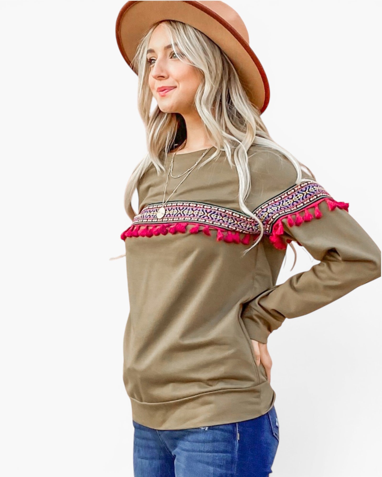 A woman wearing a brown wide-brim hat and a brown long-sleeve top with colorful geometric embroidery across the chest and sleeves, finished with bright pink tassels, paired with blue jeans against a white background.