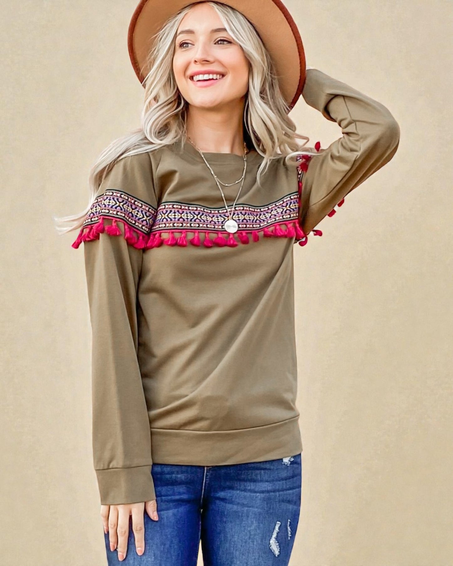 Woman wearing a beige sweater with red tassel details, blue jeans, and a brown hat on a beige background