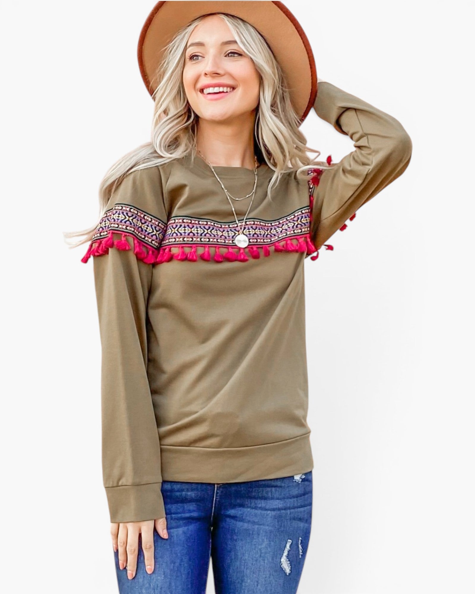 A woman wearing a long-sleeve olive green top with a colorful embroidered band and bright pink fringe trim across the chest, paired with blue jeans and a brown wide-brim hat, against a light gray studio background.