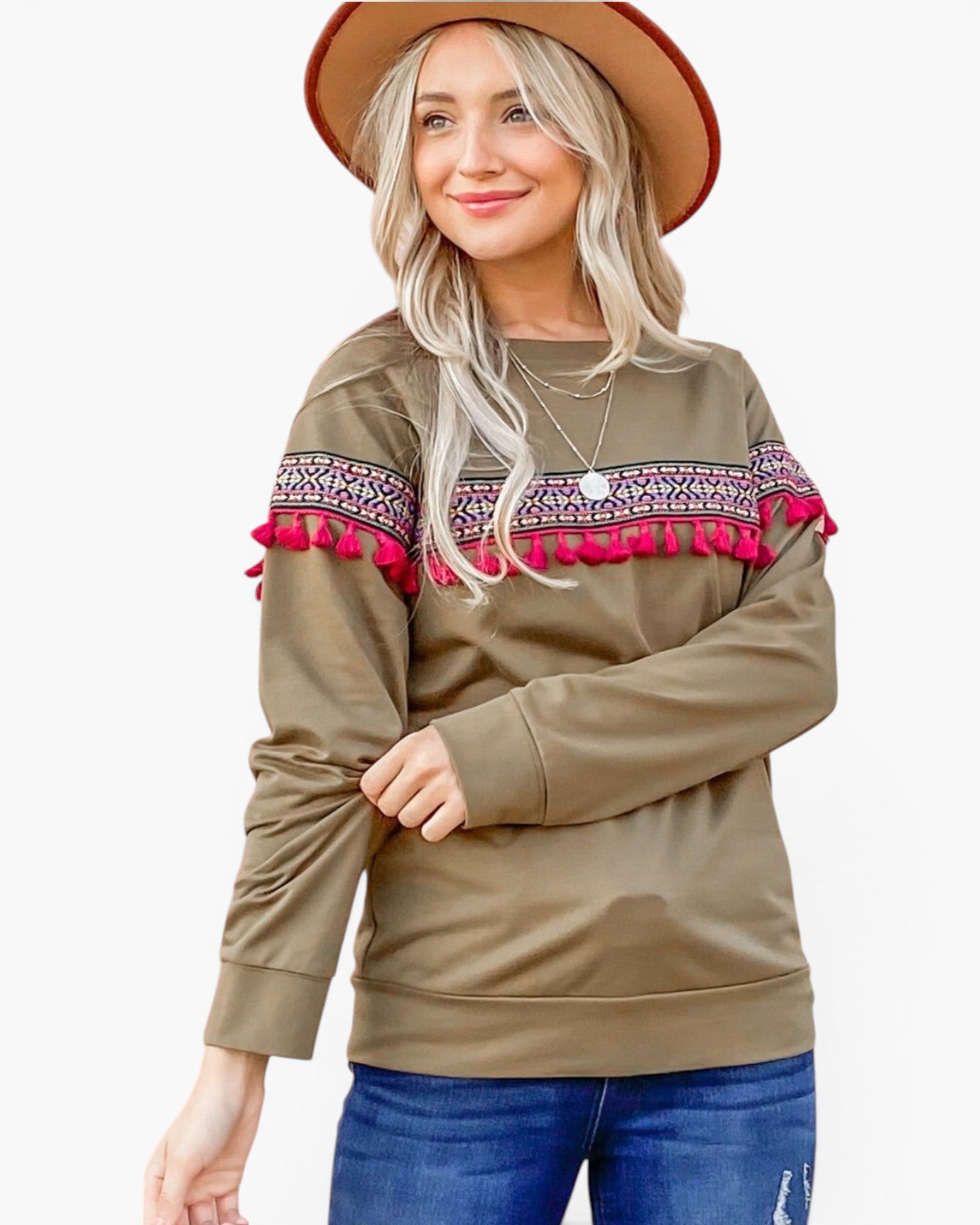 A woman wearing a brownish-olive long-sleeve top with a colorful geometric pattern and bright pink tassel trim across the shoulders, paired with blue jeans and a tan hat, against a plain white background.