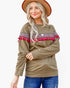 A woman wearing a brownish-olive long-sleeve top with a colorful geometric pattern and bright pink tassel trim across the shoulders, paired with blue jeans and a tan hat, against a plain white background.