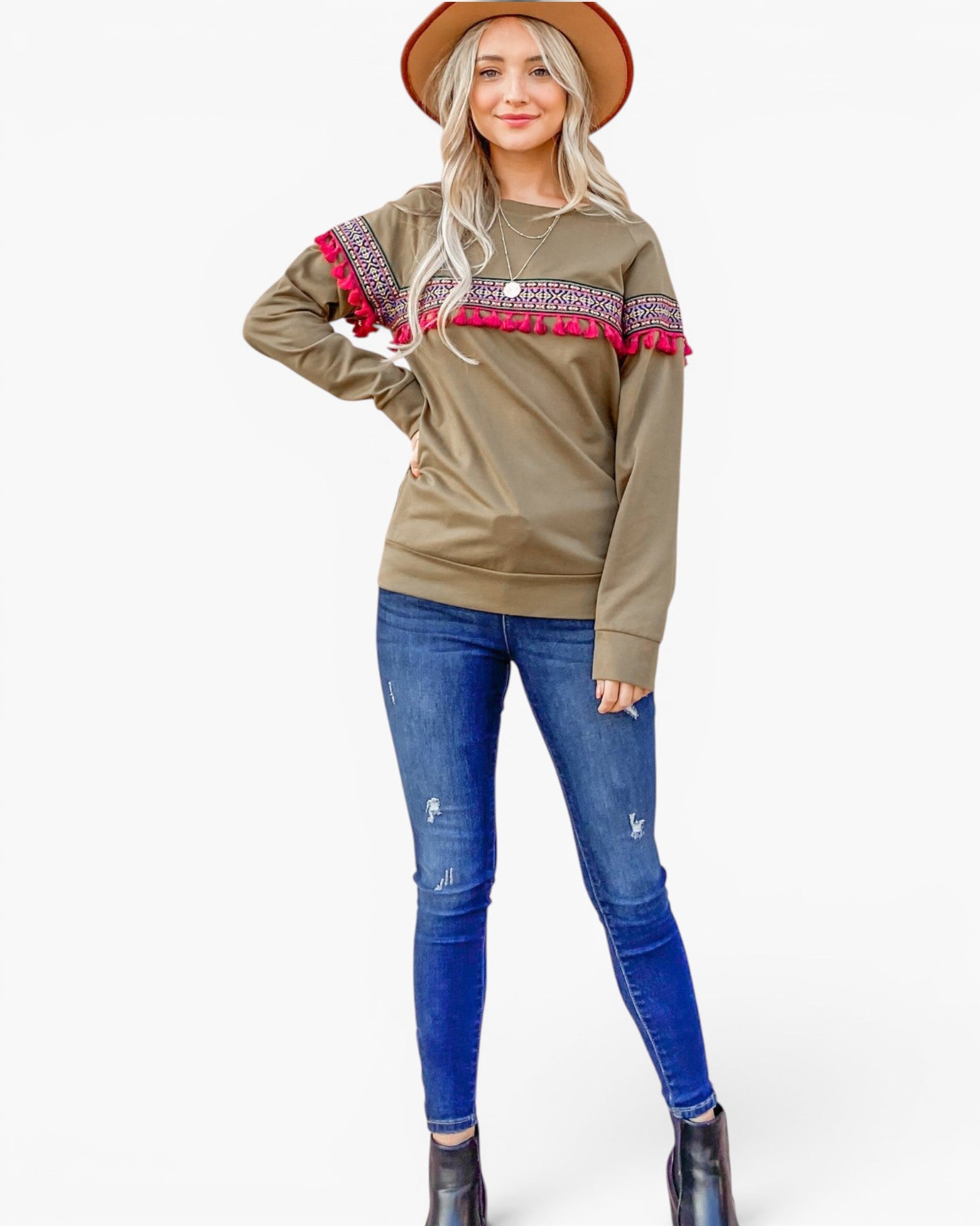 A woman modeling a brownish-olive long-sleeve sweatshirt with colorful patterned trim and pink pom-poms across the chest, paired with blue jeans and a brown hat, against a light gray studio background.