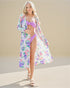 Woman in a floral cover-up and pink bikini standing on sand with a beige background