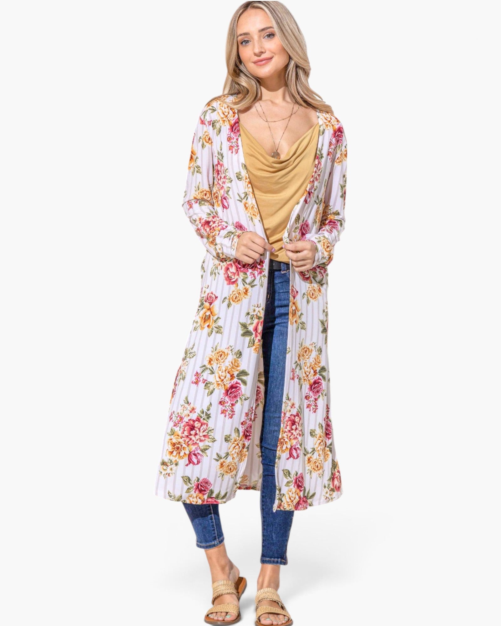 A woman modeling a long white open-front cardigan with a colorful floral print, worn over a mustard cowl-neck top and blue jeans, against a plain white background.