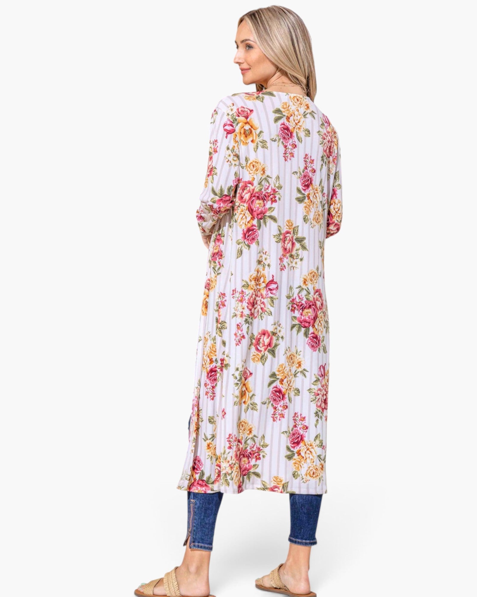 A woman shown from the back wearing a long-sleeve floral tunic dress with vertical pleats, paired with blue jeans and tan slide sandals, against a plain white background.