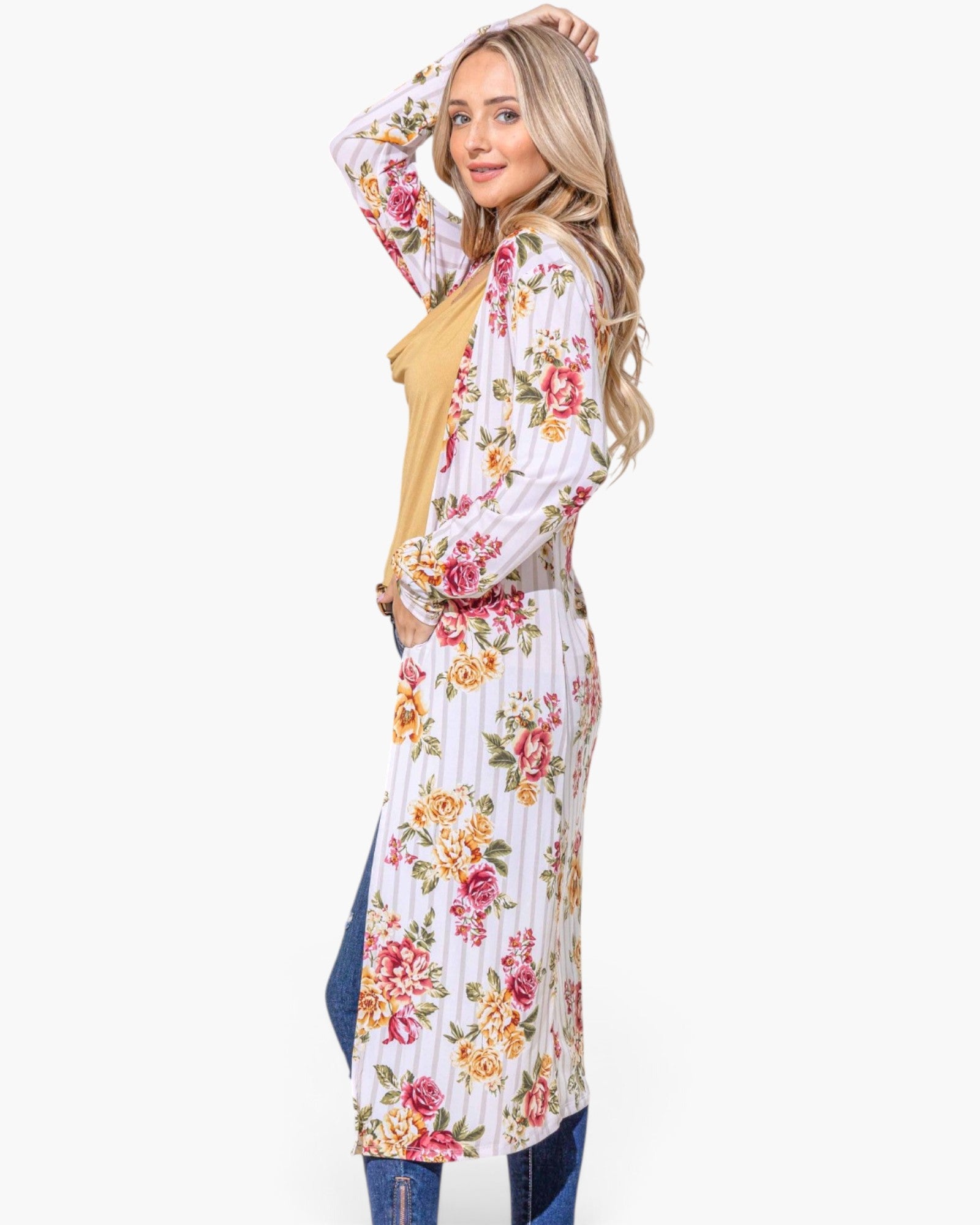 A woman modeling a long-sleeve maxi dress with a white base, vertical pink and yellow floral stripes, and a mustard yellow tie belt, styled with blue jeans against a light gray background.