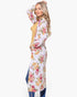 A woman modeling a long-sleeve maxi dress with a white base, vertical pink and yellow floral stripes, and a mustard yellow tie belt, styled with blue jeans against a light gray background.