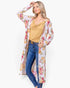 A woman modeling a long-sleeve, open-front floral kimono cardigan over a mustard yellow top and blue jeans, photographed against a plain light gray studio background.