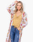 A woman modeling a long-sleeve, open-front floral kimono cardigan over a mustard yellow top and blue jeans, against a plain light gray background.