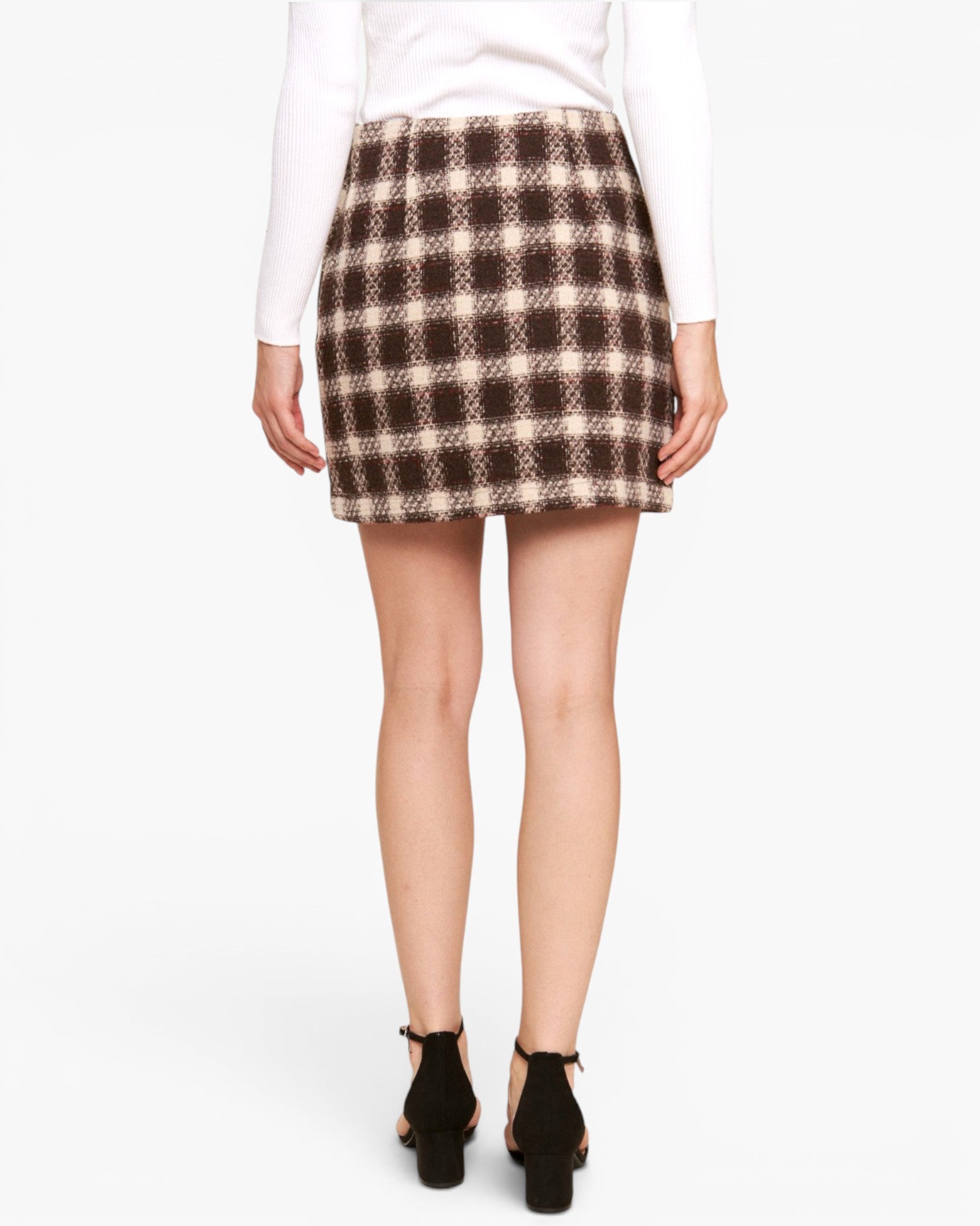 A woman shown from the back wearing a brown and beige plaid mini skirt, a white long-sleeve top, and black ankle-strap heels against a plain white studio background.