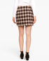 A woman shown from the back wearing a brown and beige plaid mini skirt, a white long-sleeve top, and black ankle-strap heels against a plain white studio background.