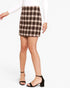 A model wearing a brown and beige plaid mini skirt with a white long-sleeve top and black strappy heels, shown from the waist down on a light gray background.