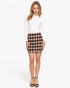 A woman modeling a white long-sleeve mock neck top and a brown and beige plaid mini skirt, paired with black strappy high-heeled sandals, against a plain white studio background.
