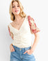 A woman modeling a cream-colored textured top with short puff sleeves featuring a red and beige floral print, paired with blue jeans against a light gray background.