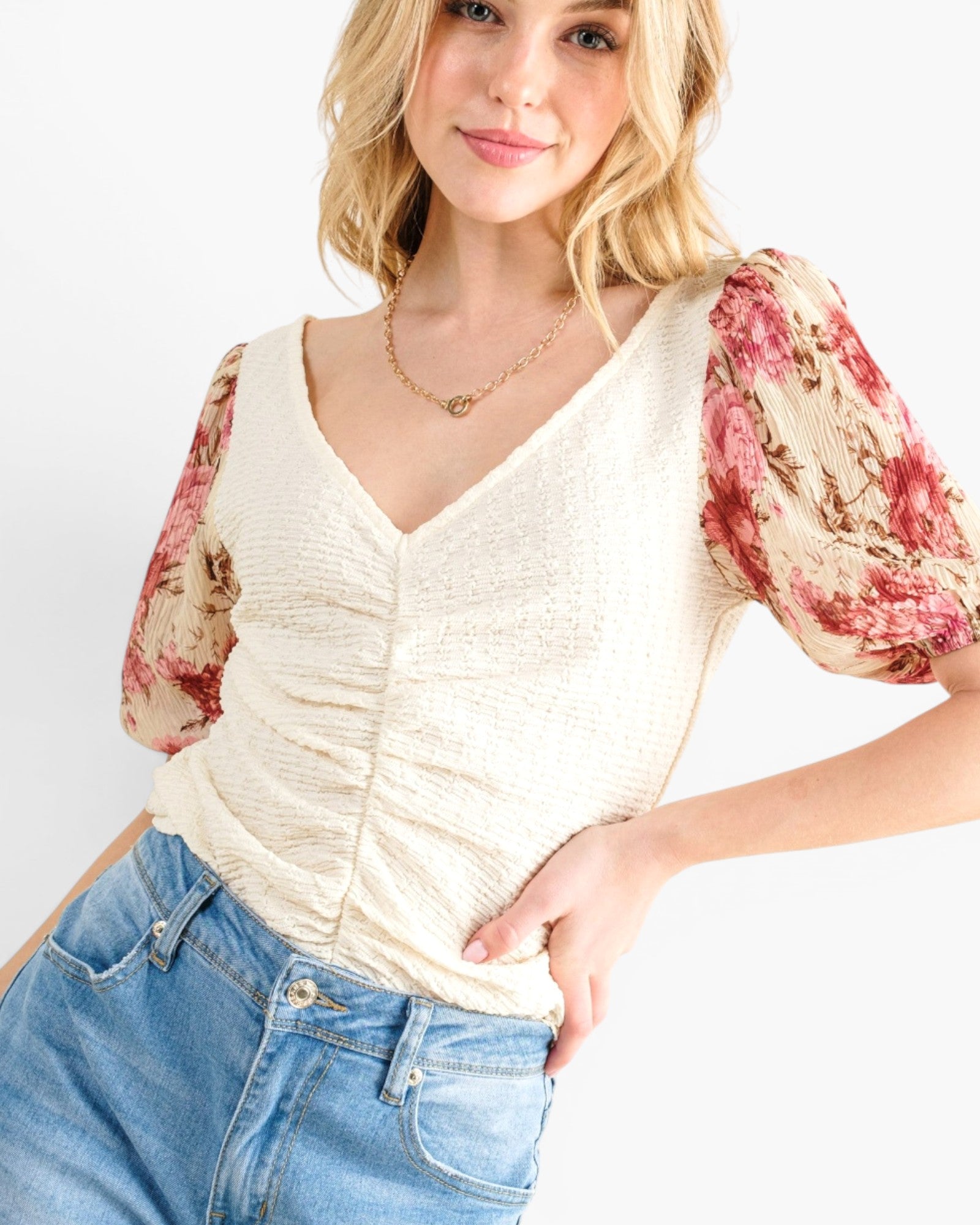 A woman wearing a cream-colored textured top with short puff sleeves featuring a red and beige floral print, paired with blue jeans against a plain white background.