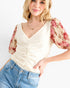 A woman wearing a cream-colored textured top with short puff sleeves featuring a red and beige floral print, paired with blue jeans against a plain white background.