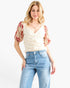 A woman modeling a cream-colored textured top with red floral puff sleeves, paired with light blue jeans, against a plain white background.