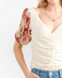 A close-up of a woman wearing a cream-colored textured top with a floral puff sleeve in red and beige, paired with blue jeans and a gold necklace.