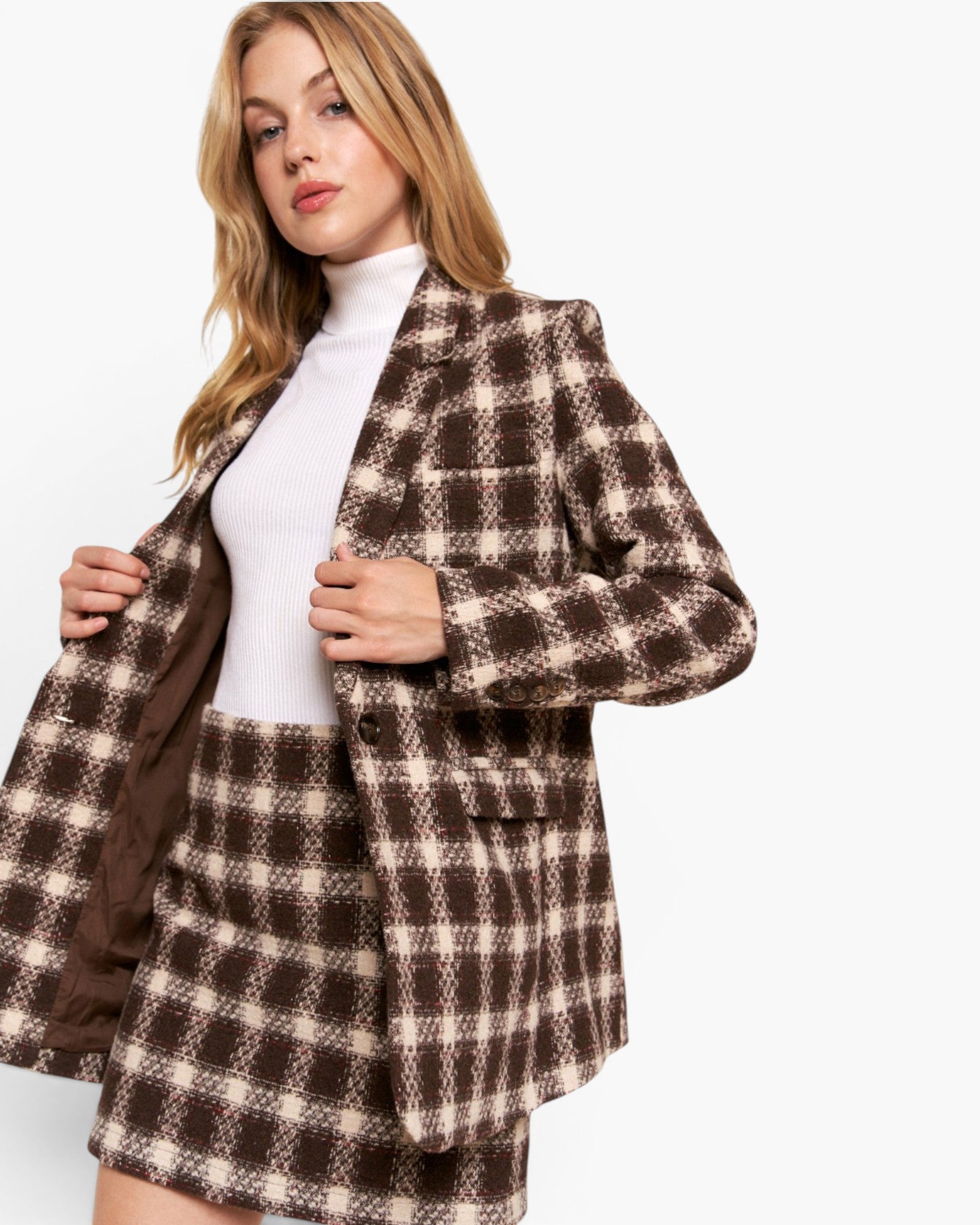 A woman modeling a brown and beige plaid blazer and matching mini skirt over a white ribbed turtleneck, photographed against a clean white studio background.