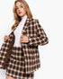 A woman modeling a brown and beige plaid blazer and matching mini skirt over a white ribbed turtleneck, photographed against a clean white studio background.