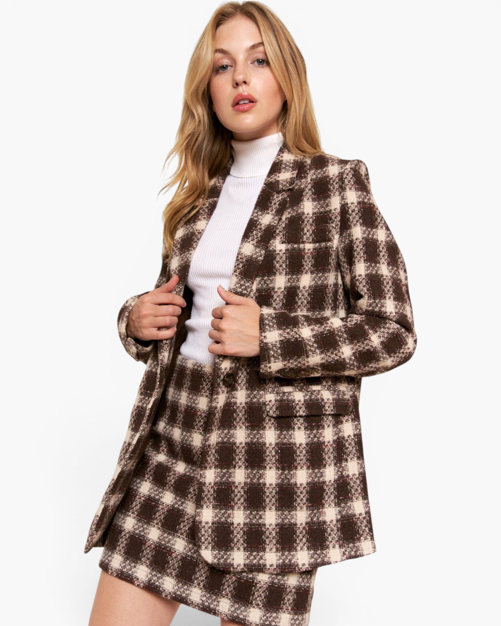 A woman modeling a brown and beige plaid blazer and matching mini skirt over a white turtleneck, set against a plain light gray studio background.