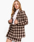 A woman modeling a brown and beige plaid blazer and matching mini skirt over a white turtleneck, set against a plain light gray studio background.