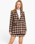 A woman modeling a brown and beige plaid blazer and matching mini skirt set over a white mock neck top, photographed against a clean white studio background.