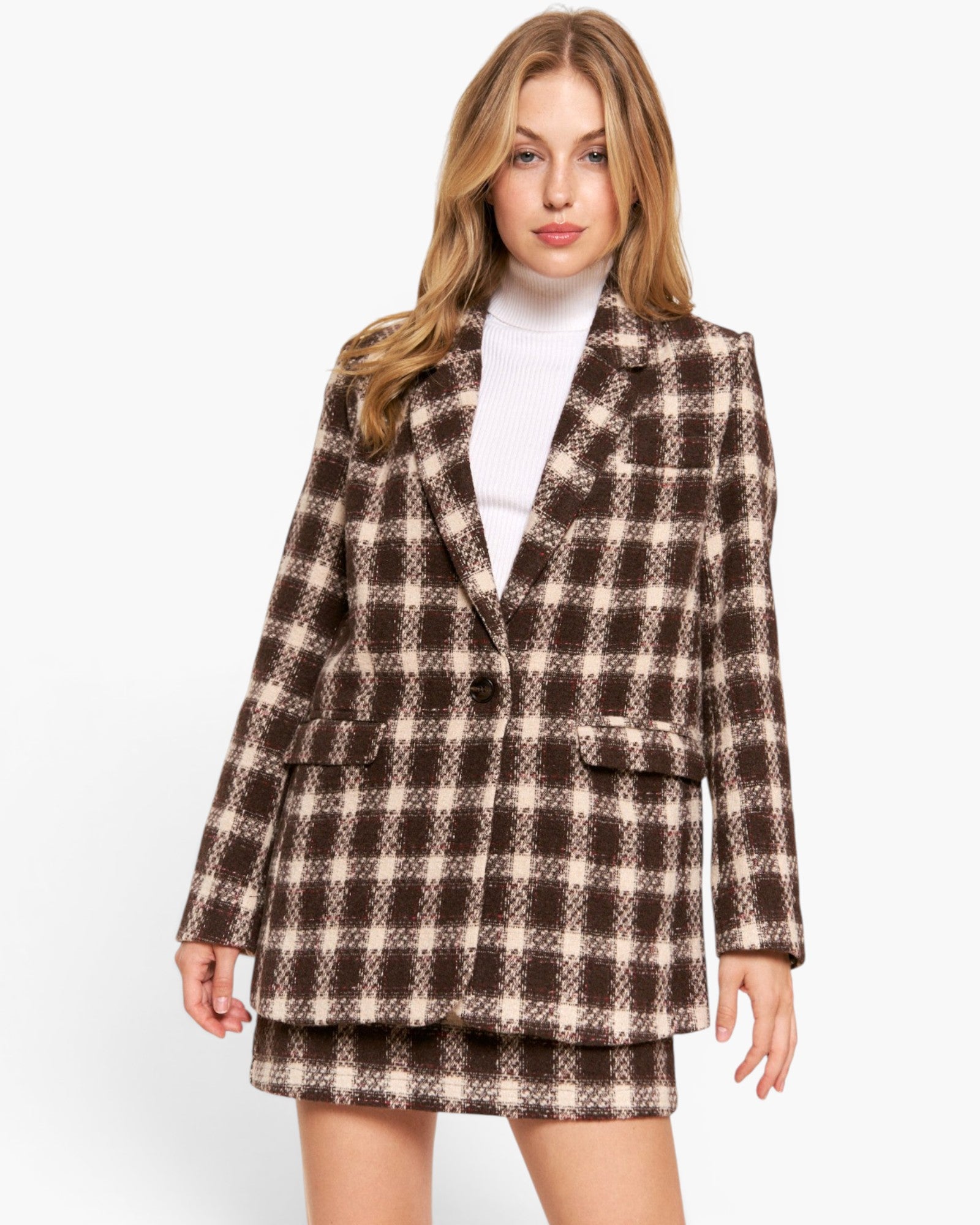 A woman modeling a brown and beige plaid blazer and matching mini skirt over a white turtleneck, shot against a plain white studio background.