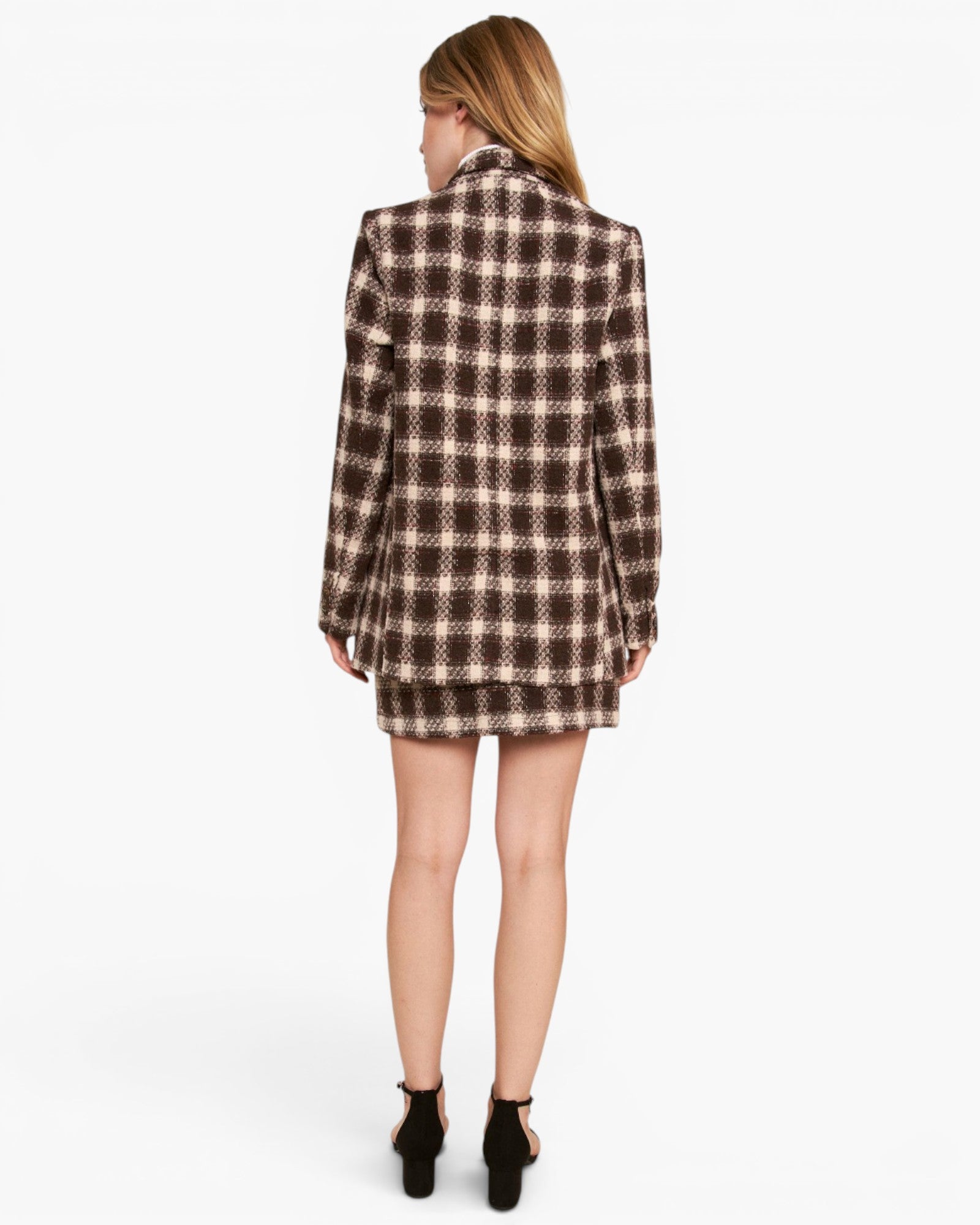 A woman viewed from behind wearing a brown and beige plaid blazer and matching mini skirt set, paired with black ankle-strap heels, against a plain white studio background.