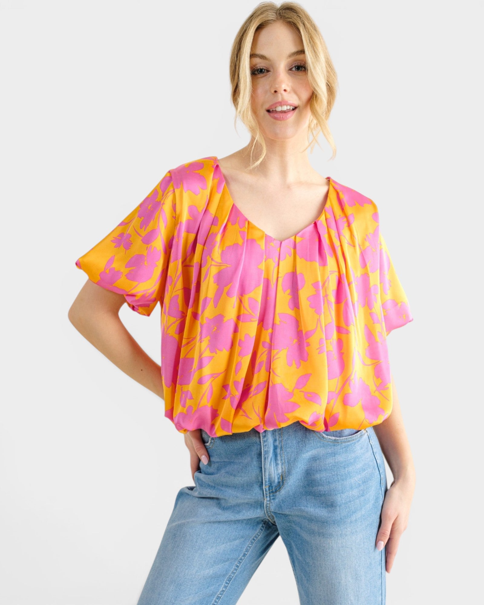 A woman wearing a short-sleeve, V-neck blouse with a bright pink and orange abstract pattern, paired with light blue jeans, against a light gray studio background.